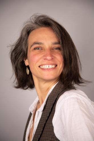 Anne Corthay, une femme aux cheveux mi-longs, portant une chemise blanche et un gilet sans manches à motifs, sur fond neutre.